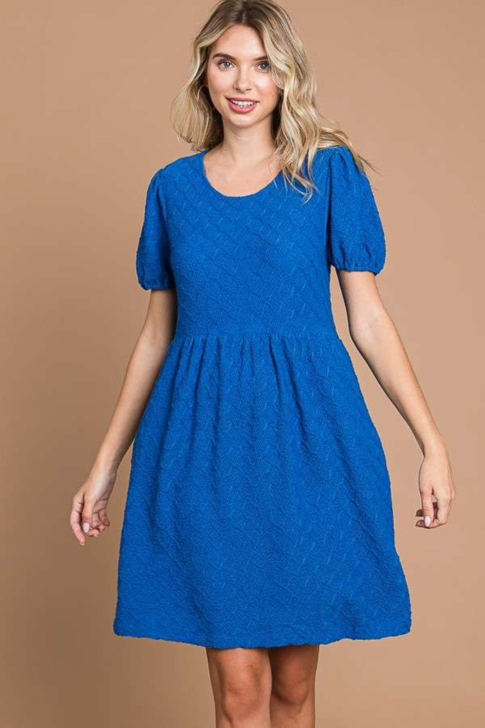 Against a plain brown backdrop, an individual is styled in a Culture Code Texture Round Neck Short Sleeve Dress with Pockets, exuding elegance and flair with its blue textured fabric and chic short puff sleeves.