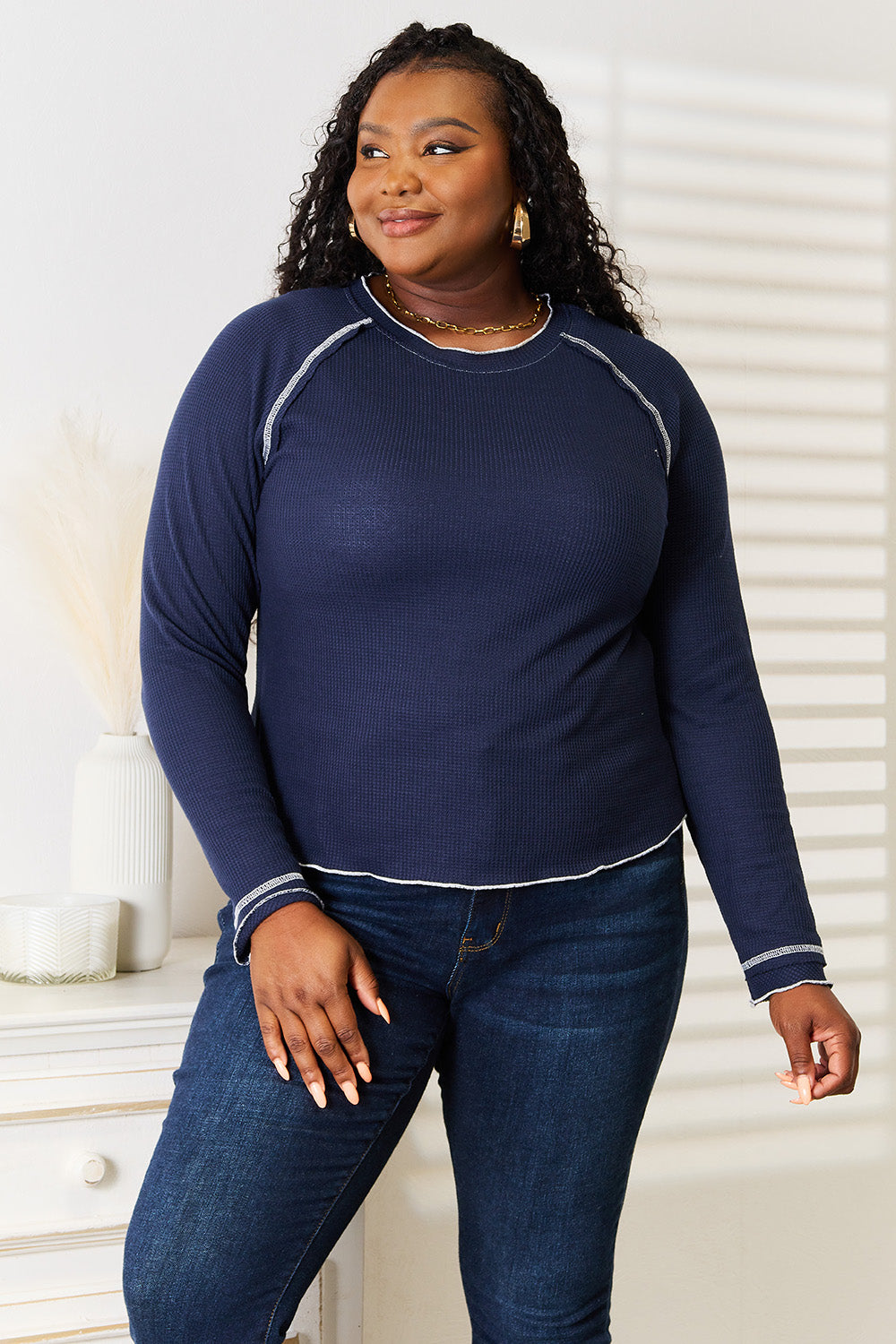 Person with long braided hair wearing a Basic Bae Long Raglan Sleeve Round Neck Top in navy blue and denim jeans, standing and smiling indoors.