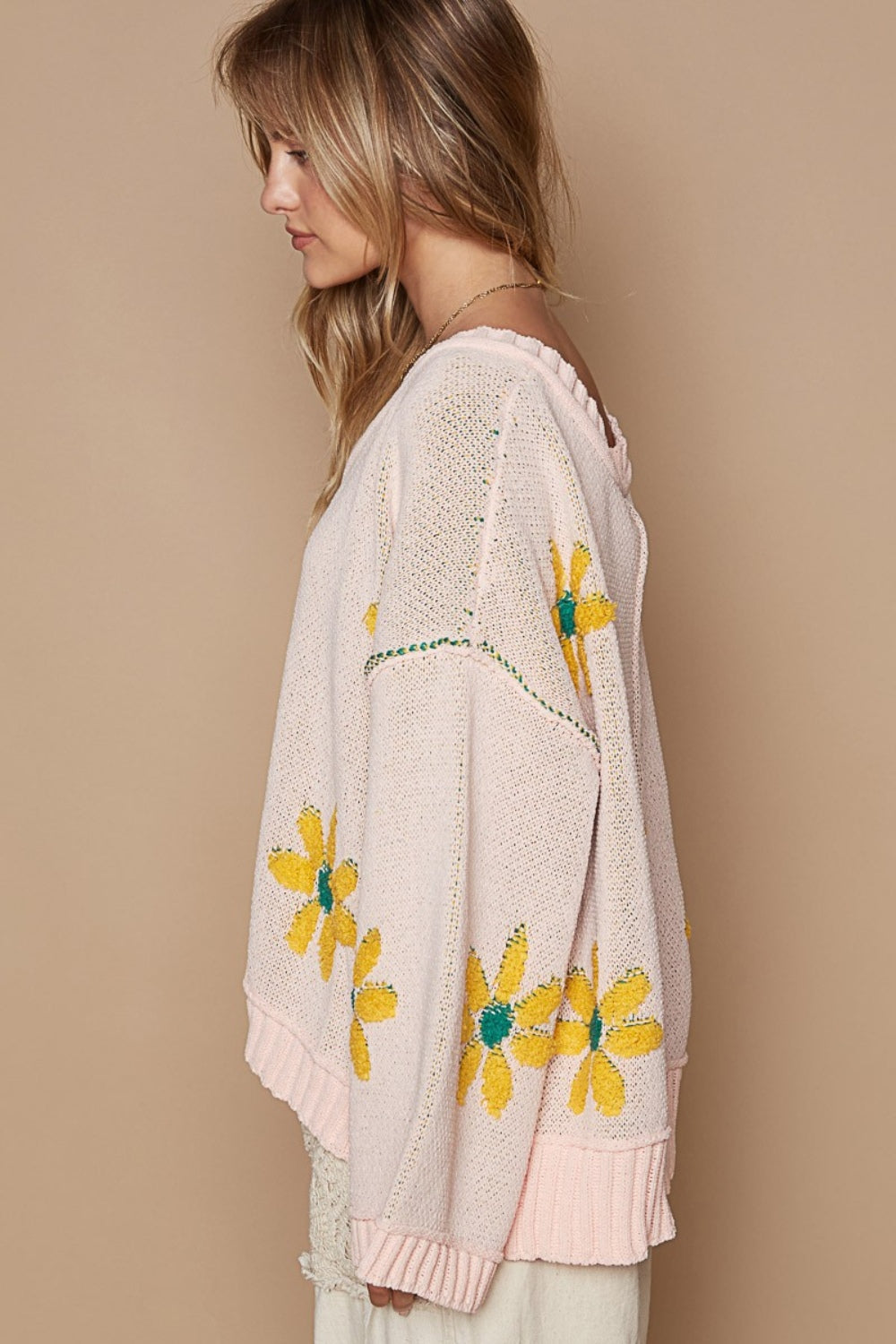 A woman with curly hair smiles while wearing a cozy POL V-Neck Floral Pattern Chenille Sweater in pink, adorned with yellow flower patterns, and holding a netted skirt in an outdoor setting.