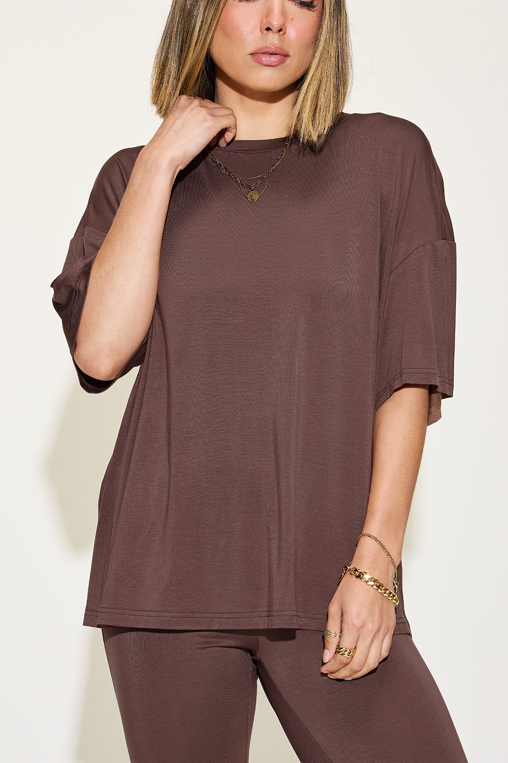 A woman with long, blonde hair wears the Basic Bae Full Size Bamboo Drop Shoulder T-Shirt and Flare Pants Set in black, paired with brown heeled sandals and layered necklaces. She stands against a plain, light background.