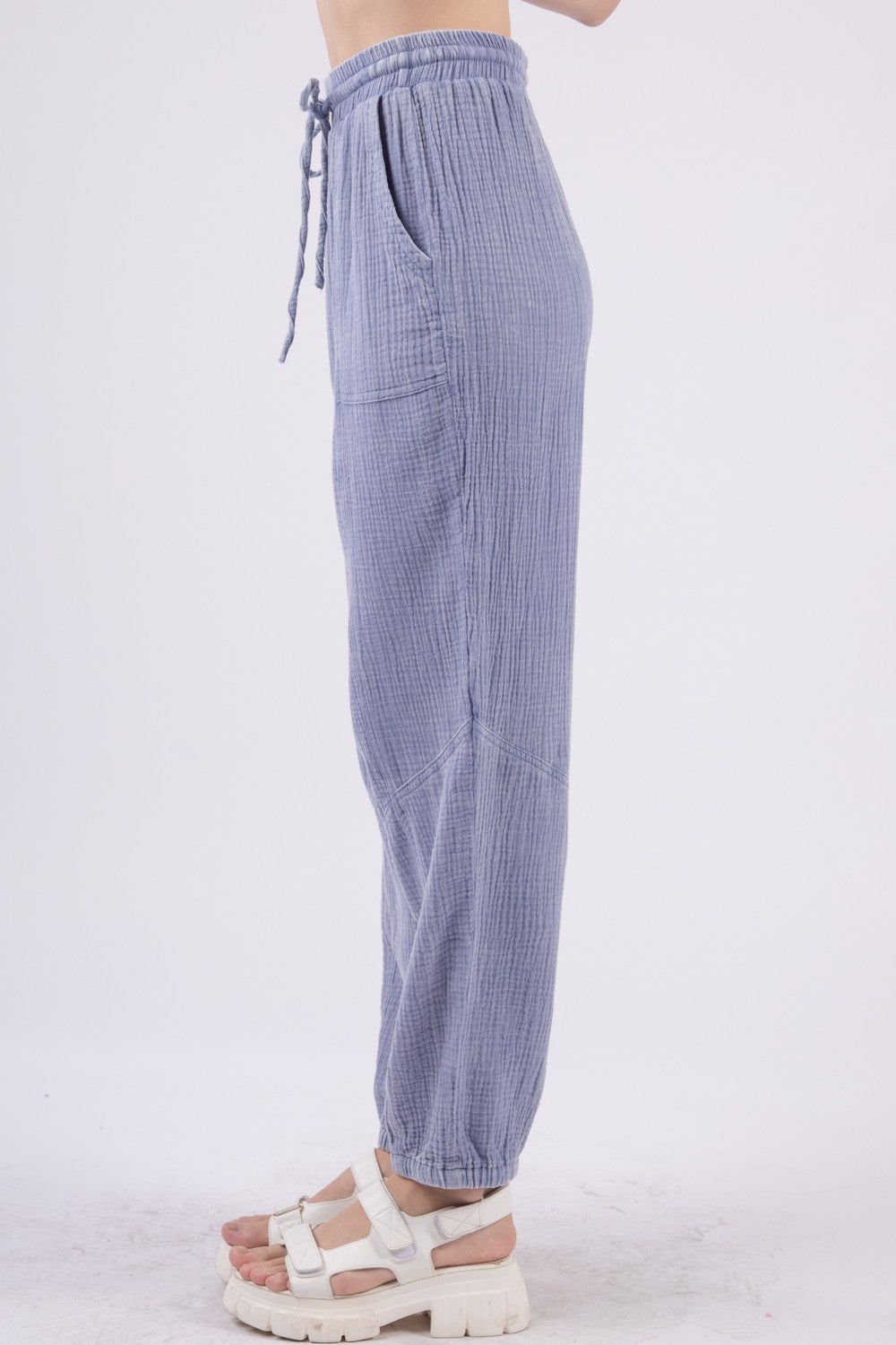 A person wearing VERY J Washed Woven Crinkle Gauze Drawstring Cargo Pants in light blue, paired with white sandals. These relaxed-fit, textured pants feature an elastic waistband and pockets, showcasing a utility design that's perfect for blending comfort and functionality against a plain background.