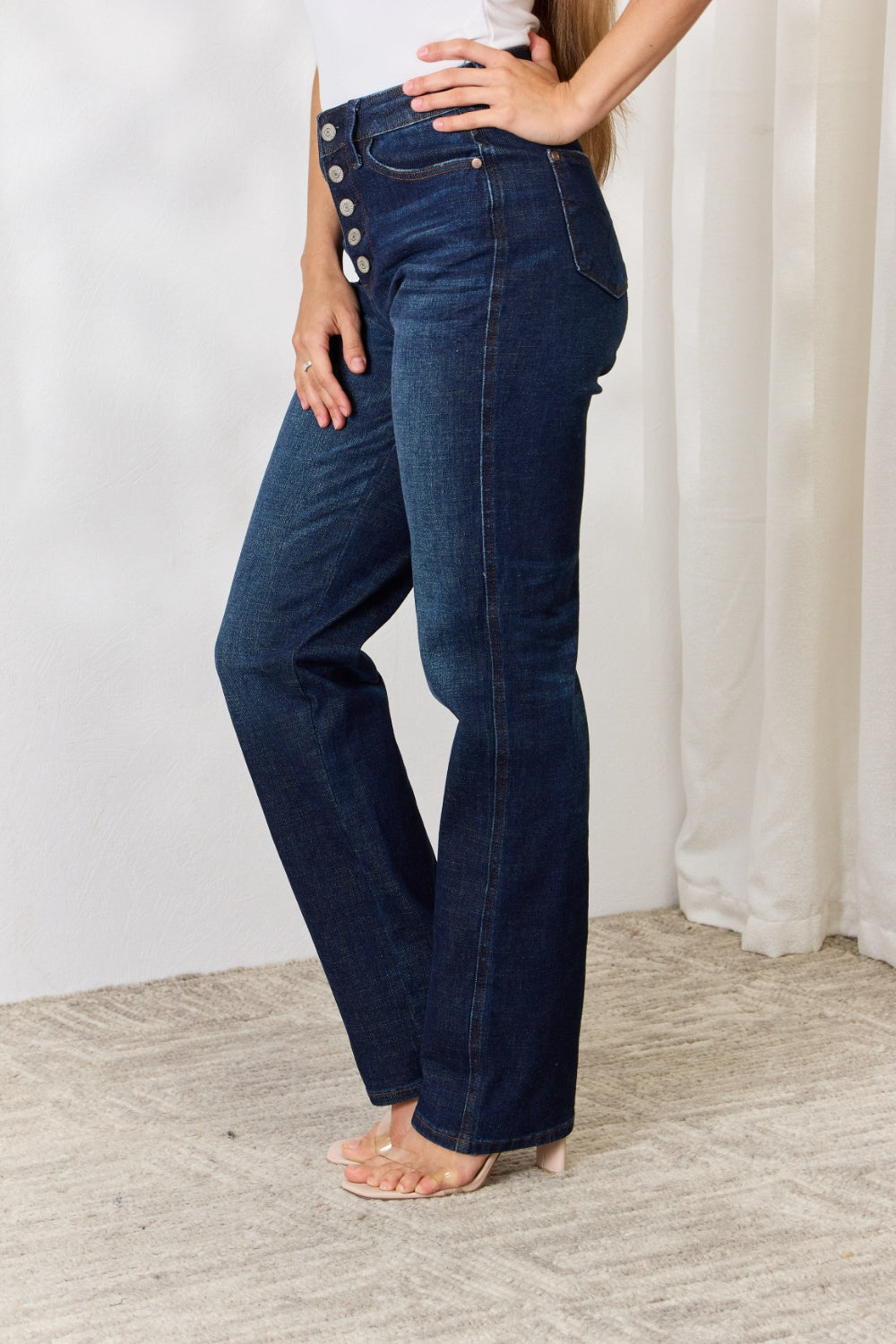 A person stands with hands in pockets, wearing the Judy Blue Full Size Button-Fly Straight Jeans, known for their high-waisted dark blue design crafted from premium lycra fabric. The jeans showcase button details and a straight leg cut, complemented by a white top and open-toed heels.