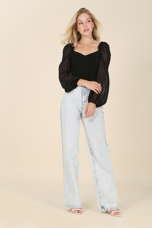 A person wearing the LS shirring sleeve bodysuit with Swiss dot mesh, featuring sheer, long puffed sleeves and a sweetheart neck, paired with light-colored jeans, stands against a plain background.