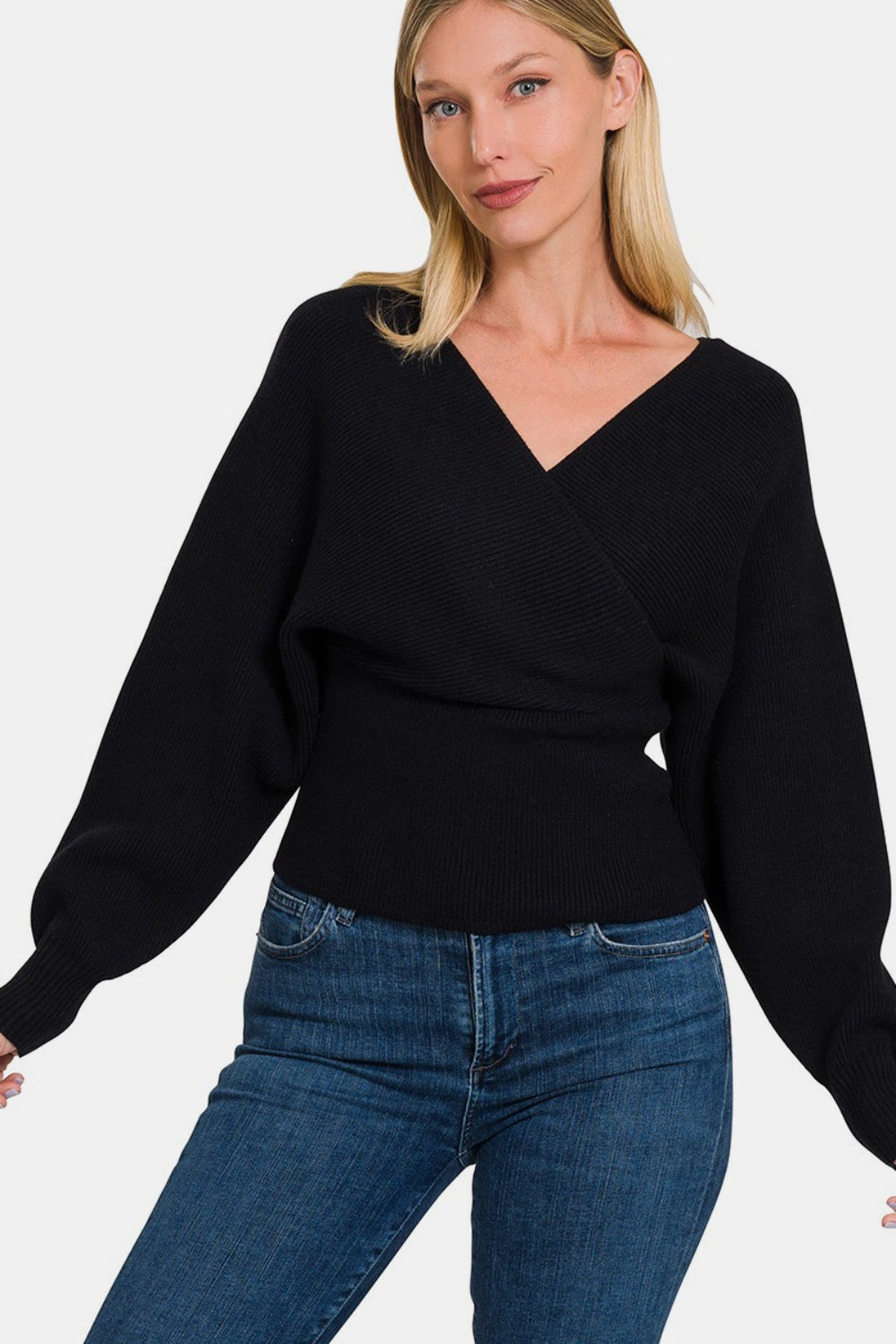 A woman stands against a plain white background, wearing the chic Zenana Cross Wrap Rib Long Sleeve Sweater in black paired with blue jeans.