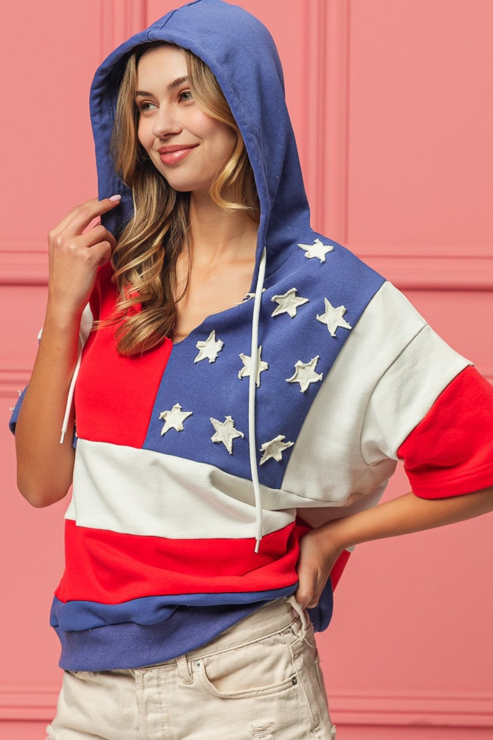 Woman wearing a BiBi American Flag Theme Hoodie, standing against a pink background.