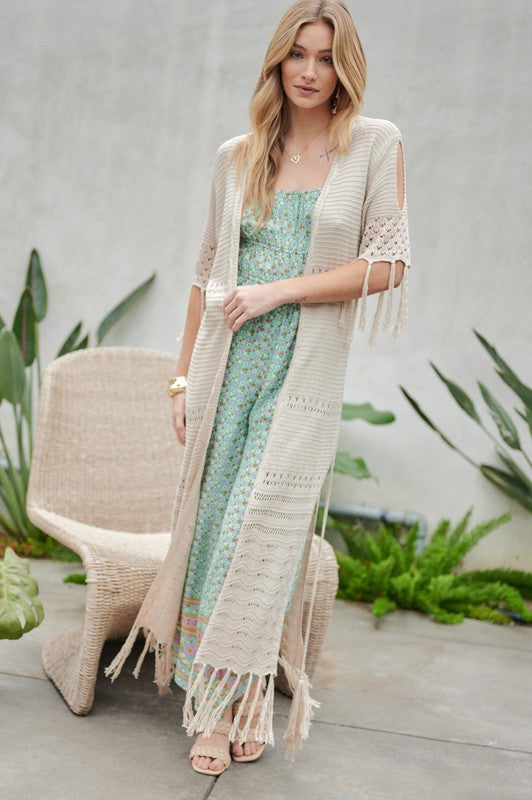 A woman in a green floral dress and a Solid Long Cardigan With Fringe exudes boho-chic vibes as she strolls on a patio, surrounded by outdoor furniture and lush plants.