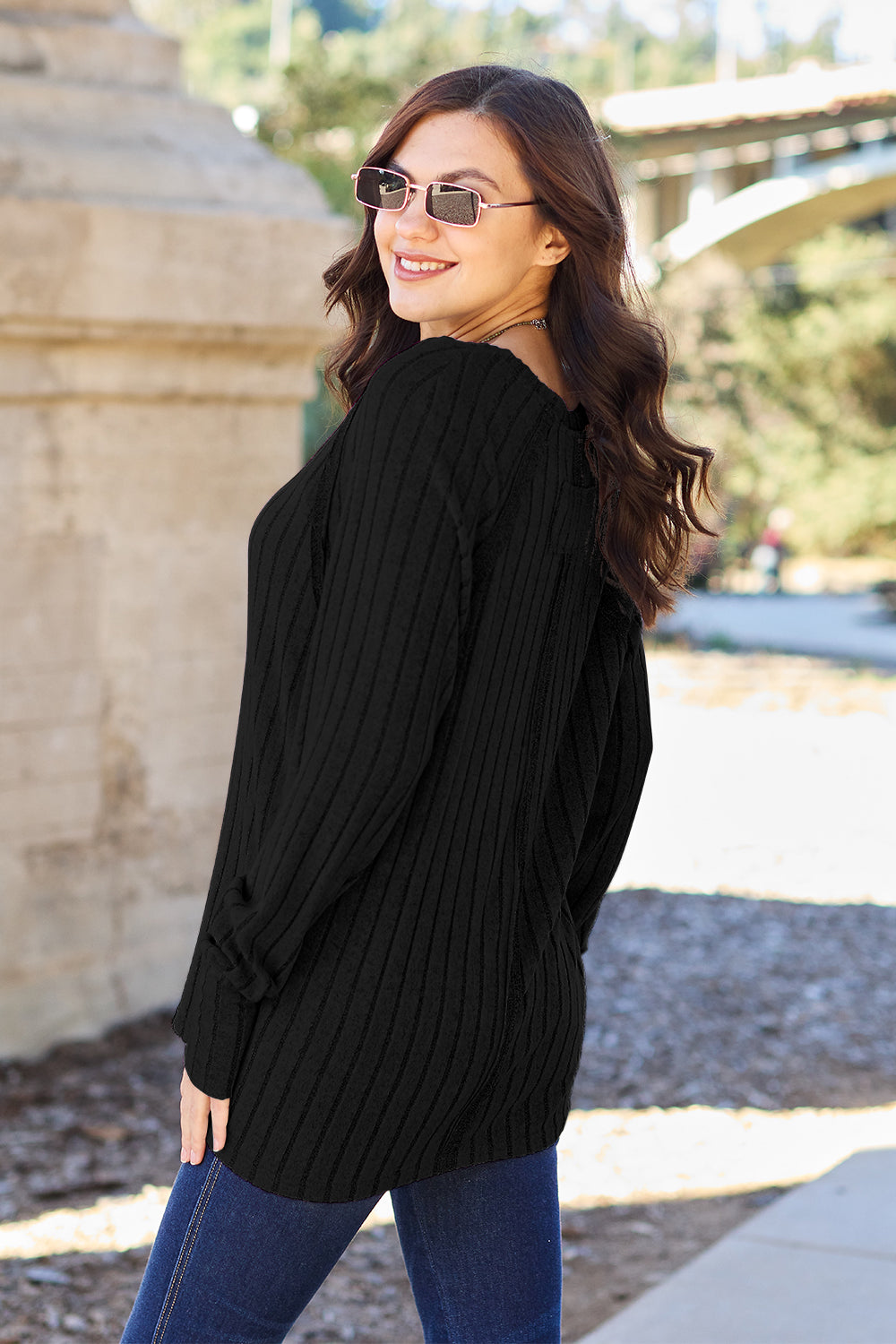 A person with long dark hair and sunglasses stands outdoors, wearing the Basic Bae Full Size Ribbed Round Neck Long Sleeve Knit Top in blue and a pair of jeans. The basic style outfit blends seamlessly with the background of trees and a bridge.