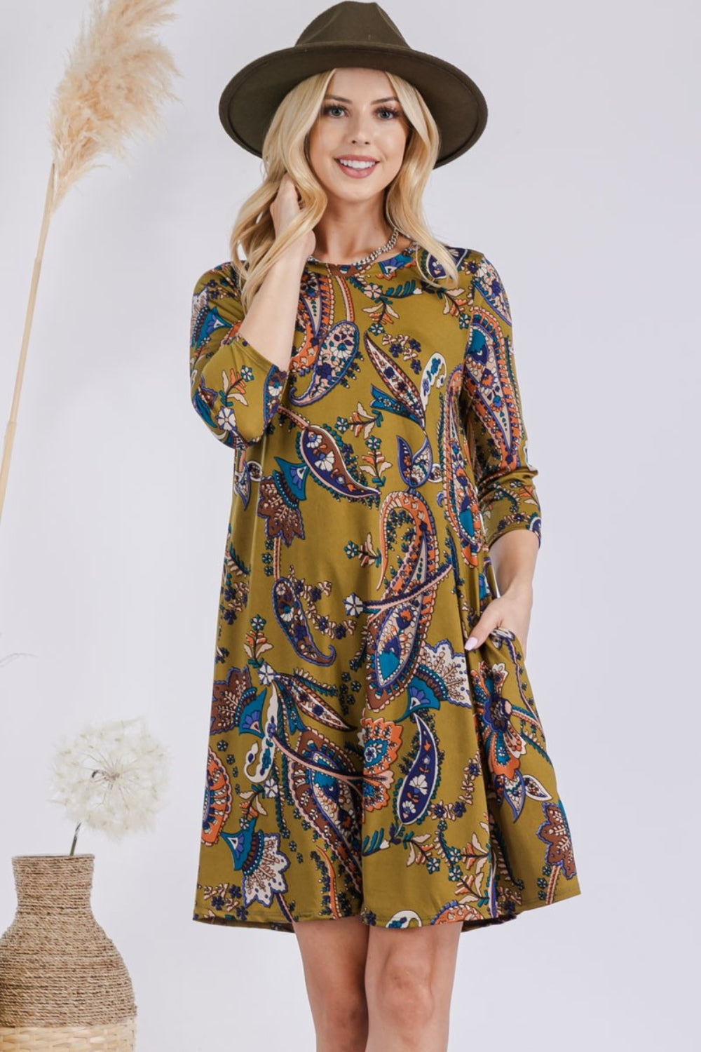 A woman wearing the chic Celeste Full Size Paisley Print Round Neck Dress with Pockets and a stylish hat stands beside decorative vases, smiling at the camera.