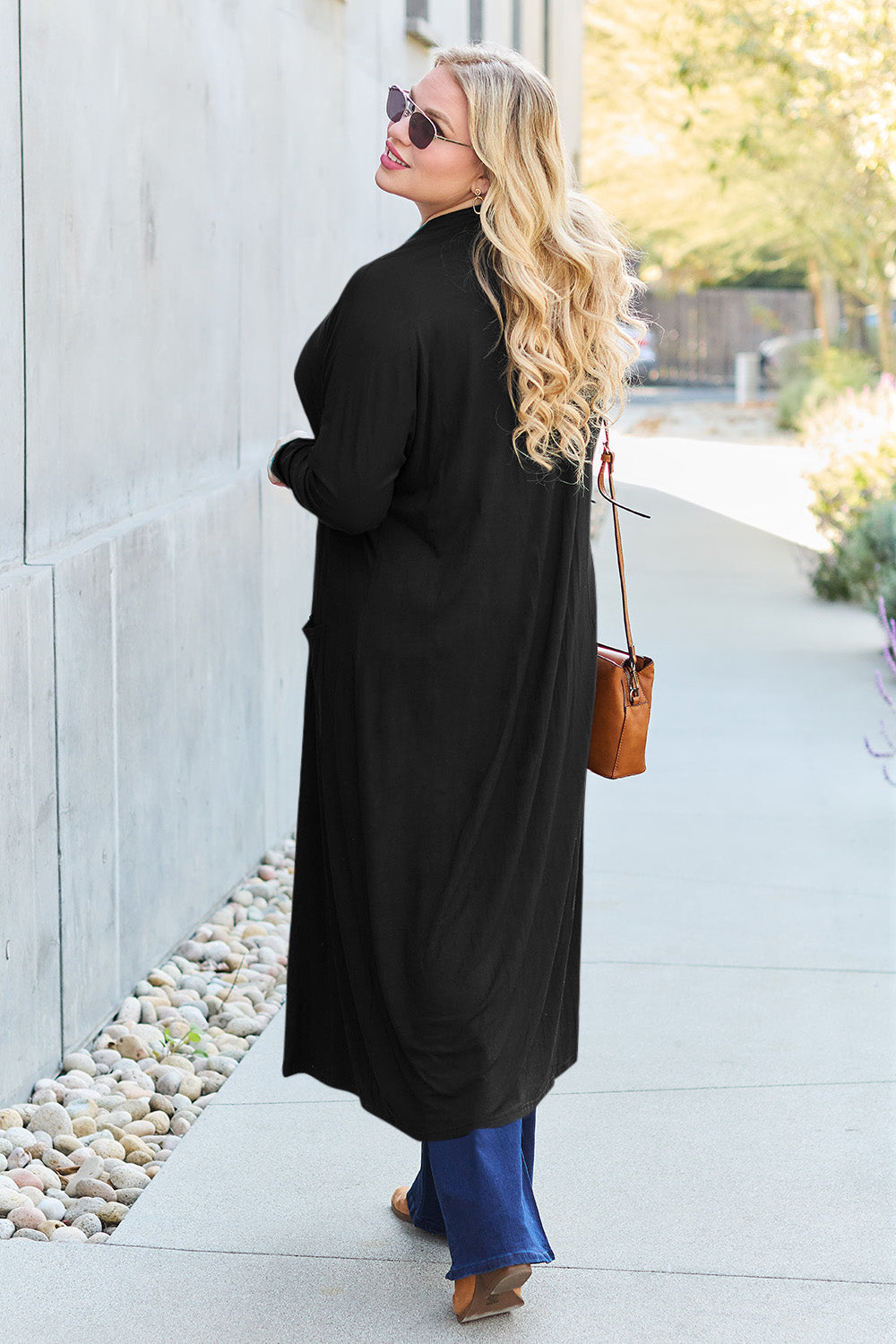 A woman stands against a concrete wall, wearing the Basic Bae Full Size Open Front Long Sleeve Cover Up made from rayon spandex, a white top, blue jeans in her perfect size, and brown shoes with her hands in her pockets. Machine wash cold for best results.