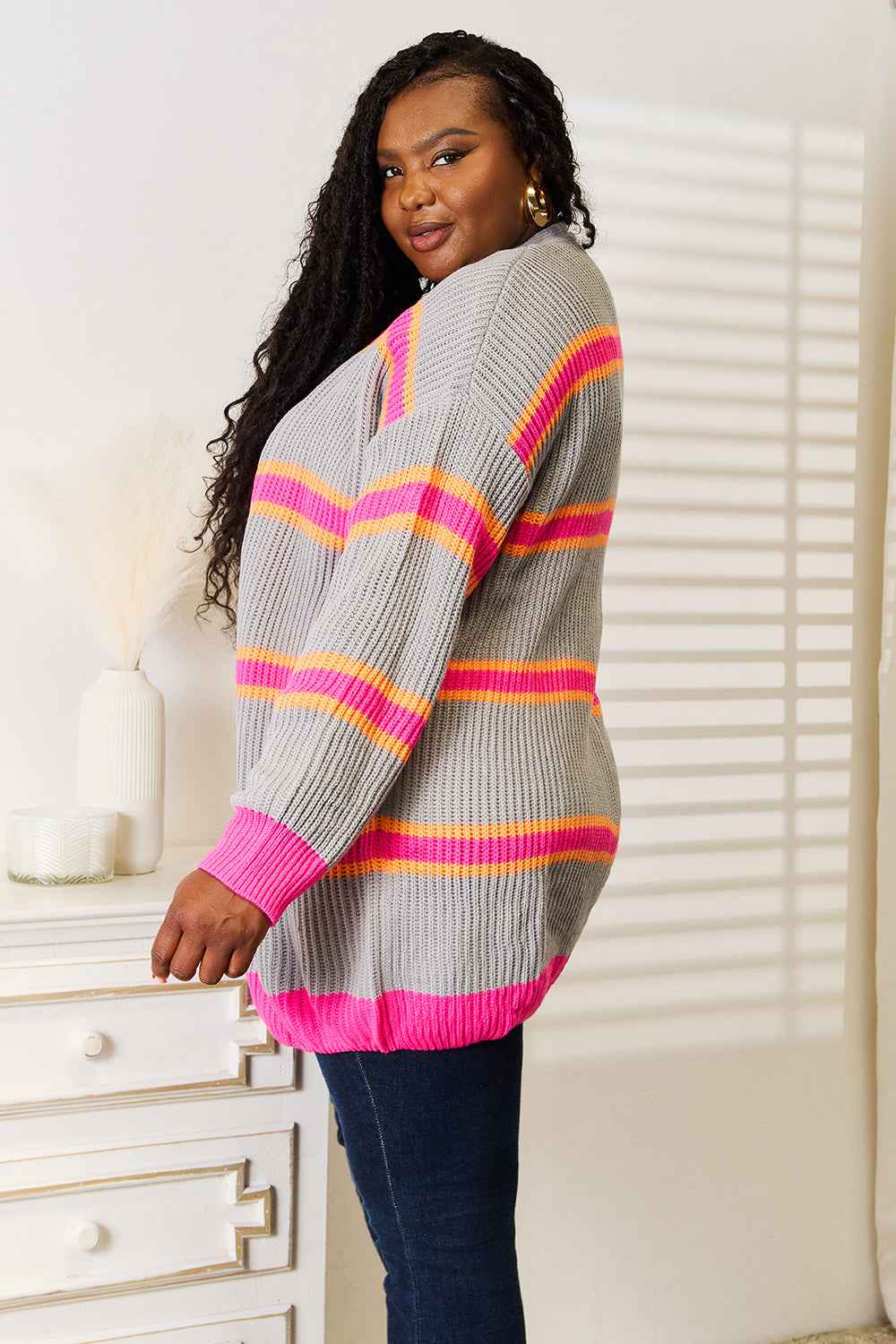 A woman stands against a white wall, smiling with one hand on her hip. She wears the Woven Right Ribbed Long Sleeve Cardigan, featuring pink and orange stripes, over a white top and dark jeans—an excellent choice for versatile layering.