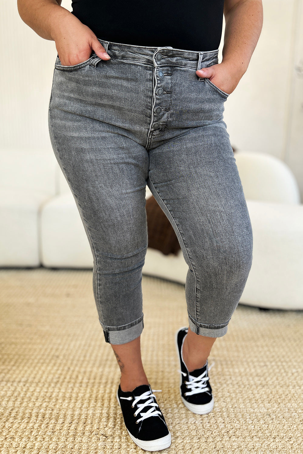 A person wearing Judy Blue Full Size Button Fly High Waist Cuffed Capris in gray, paired with a white top and white sneakers, stands on a beige carpeted floor.