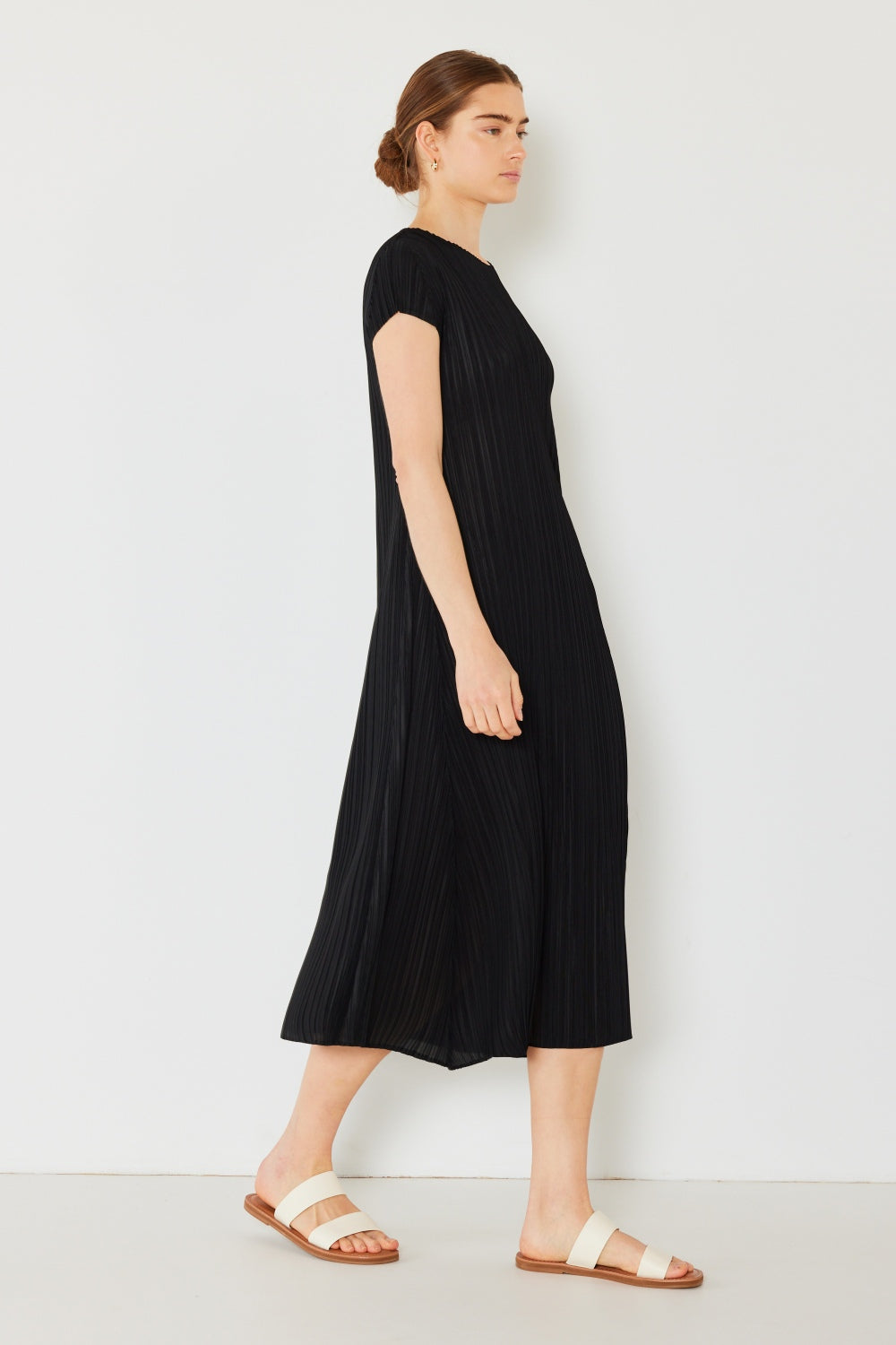 A person stands against a light wall, wearing an elegant Marina West Swim Pleated Cap Sleeve A-Line Dress in black and white sandals.