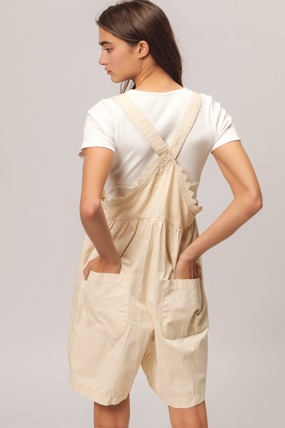 A young woman with long dark hair is sitting on a wooden stool, wearing a white t-shirt paired with the BiBi Back Patch Pocket Lace Trim Rompers—a cream-colored overall dress featuring back patch pockets. With a neutral expression set against a plain light gray background, she effortlessly embodies the perfect addition to any summer wardrobe collection.