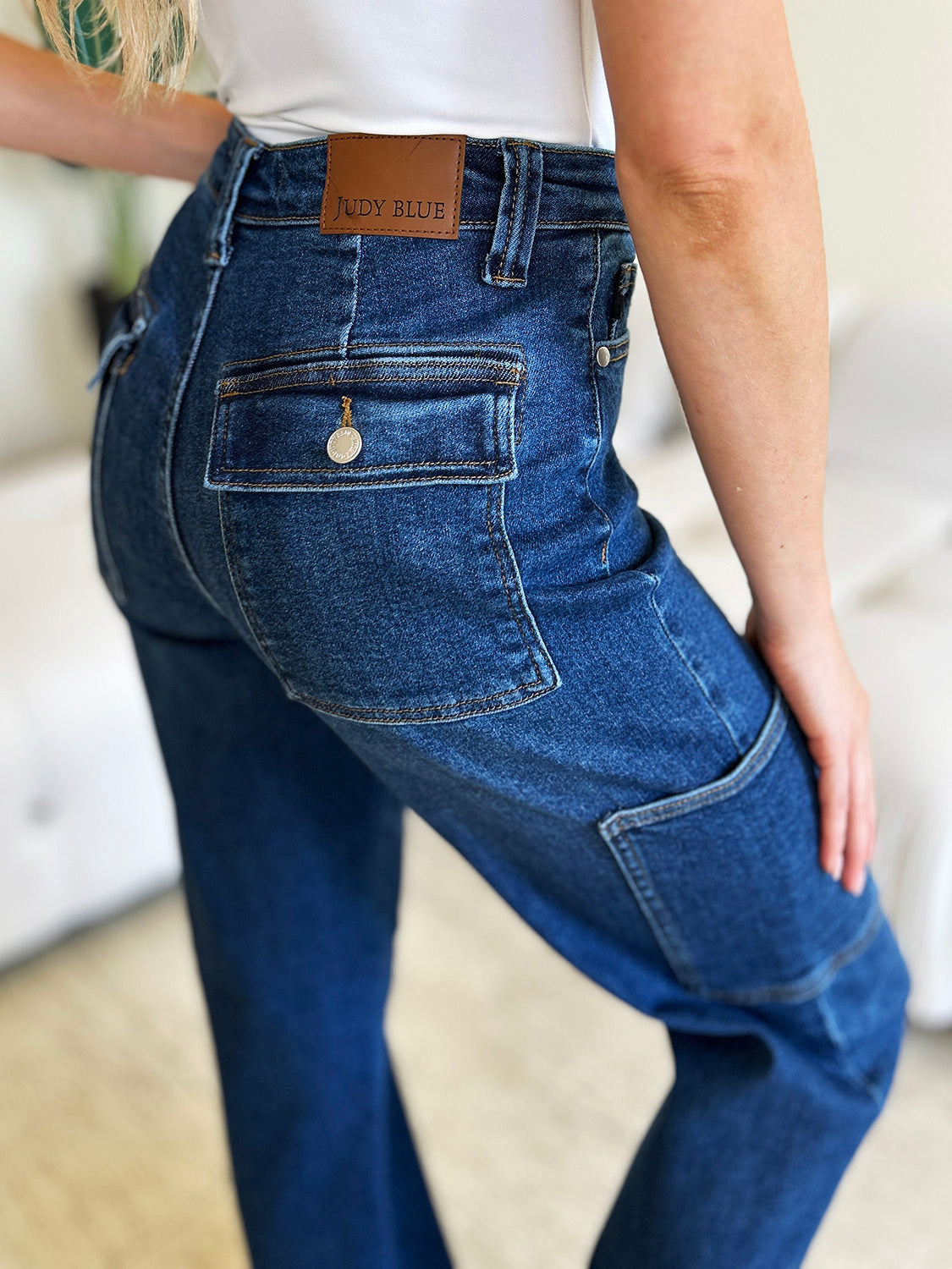 A person wearing Judy Blue Full Size High Waist Straight Cargo Jeans made of durable denim fabric and a white top walks on a wooden floor, with a white sofa and green plant in the background.