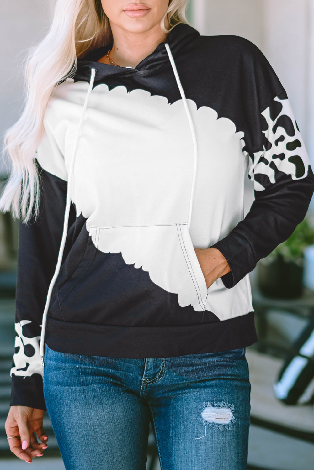 A person with long blonde hair is seen from the back wearing a Black Cow Tie Dye Print Pocketed Drawstring Pullover Hoodie and blue jeans. They are standing in an indoor space with a window and furniture in the background.