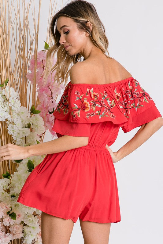 Wearing the stylish Floral Embroidered Off Shoulder Romper, a woman poses gracefully, showcasing its ruffle detail and making it an ideal addition to any summer wardrobe.