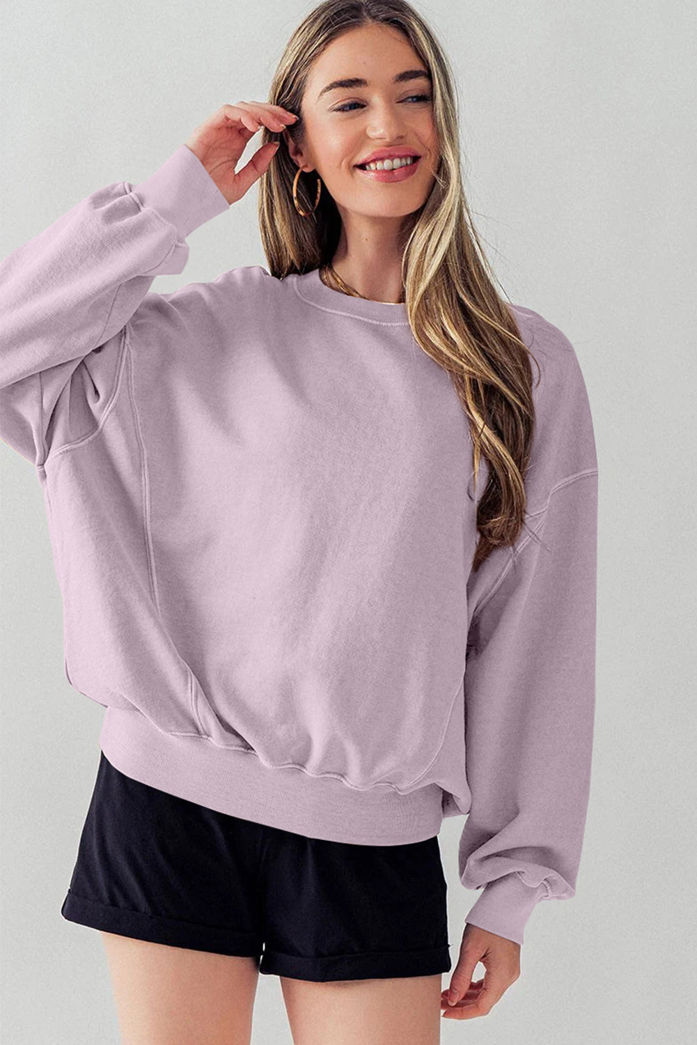 A woman facing away wears the Orchid Petal Exposed Seam Batwing Sleeve Drop Shoulder Sweatshirt and black shorts. She poses with her right arm raised, resting on her head.