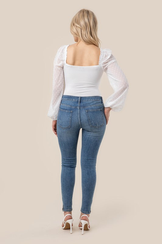 A person wearing the LS shirring sleeve bodysuit with Swiss dot mesh, featuring sheer, long puffed sleeves and a sweetheart neck, paired with light-colored jeans, stands against a plain background.