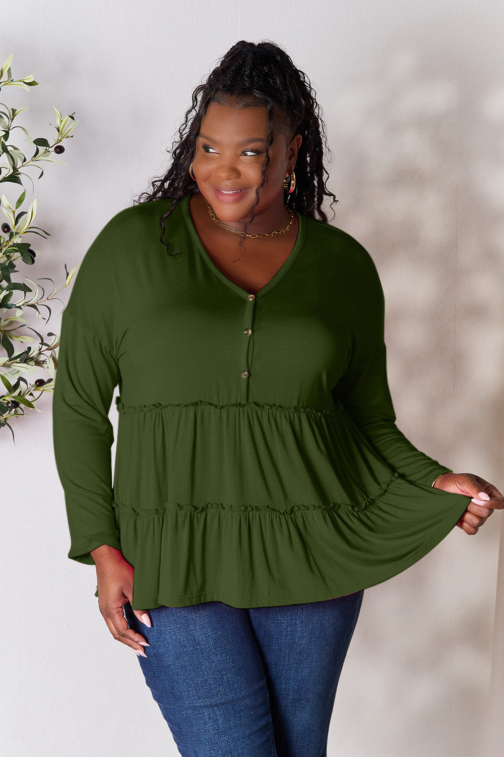 Person stands indoors near a plant, wearing the Double Take Half Button Long Sleeve Ruffle Hem Blouse in gray and blue jeans, smiling while looking to the side. The slightly stretchy fabric of the blouse adds comfort while its buttoned design offers a touch of elegance.