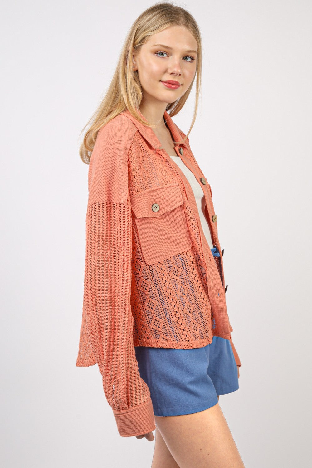 A young woman with blonde hair is seen standing against a plain background, smiling slightly. She wears the VERY J Button Up Long Sleeve Lace Shirt over a white top, paired with blue shorts.