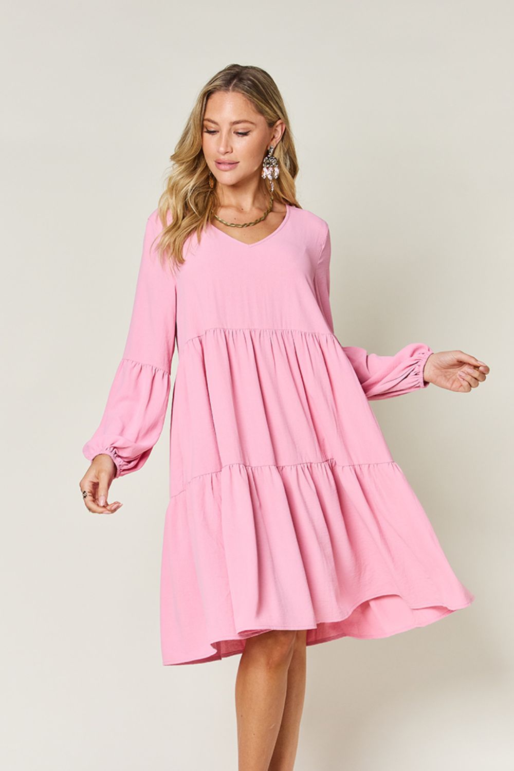 A woman stands wearing the Double Take Full Size V-Neck Balloon Sleeve Tiered Dress with Pockets in pink. Crafted from high stretch yarn, this dress combines comfort and style effortlessly. She has long, wavy hair and accessorizes with hoop earrings and a necklace. This imported garment is easy to care for – simply machine wash cold to keep it looking fresh.