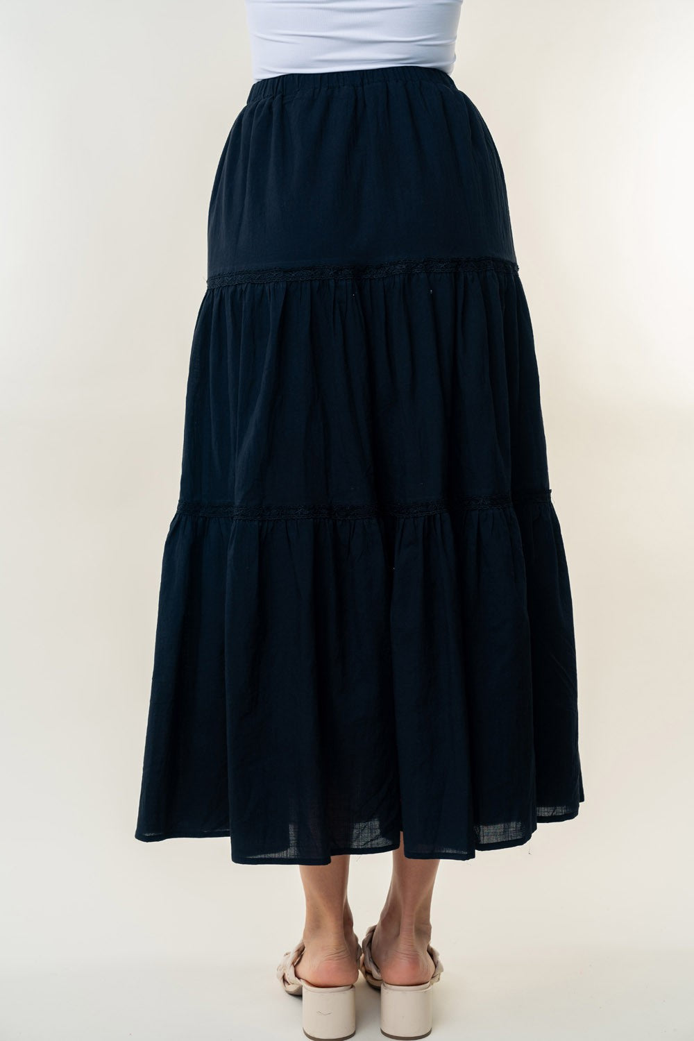 A long, White Birch High Waisted Tiered Maxi Skirt in navy blue, offering a versatile piece perfect for a bohemian look. It's worn by a person paired with white heeled sandals. The background is plain and light-colored.