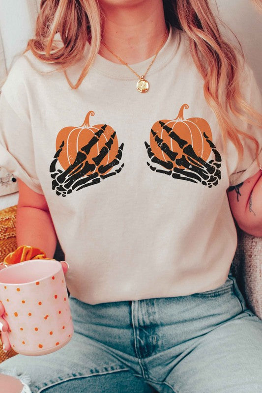 A person wearing the SKELETON HOLDING PUMPKINS Graphic Tee, which features two orange pumpkins and black skeletal hands, is sitting down. With long, wavy hair and accessorized with various bracelets and a necklace, the unisex sizing of the shirt makes it a versatile addition to any wardrobe.