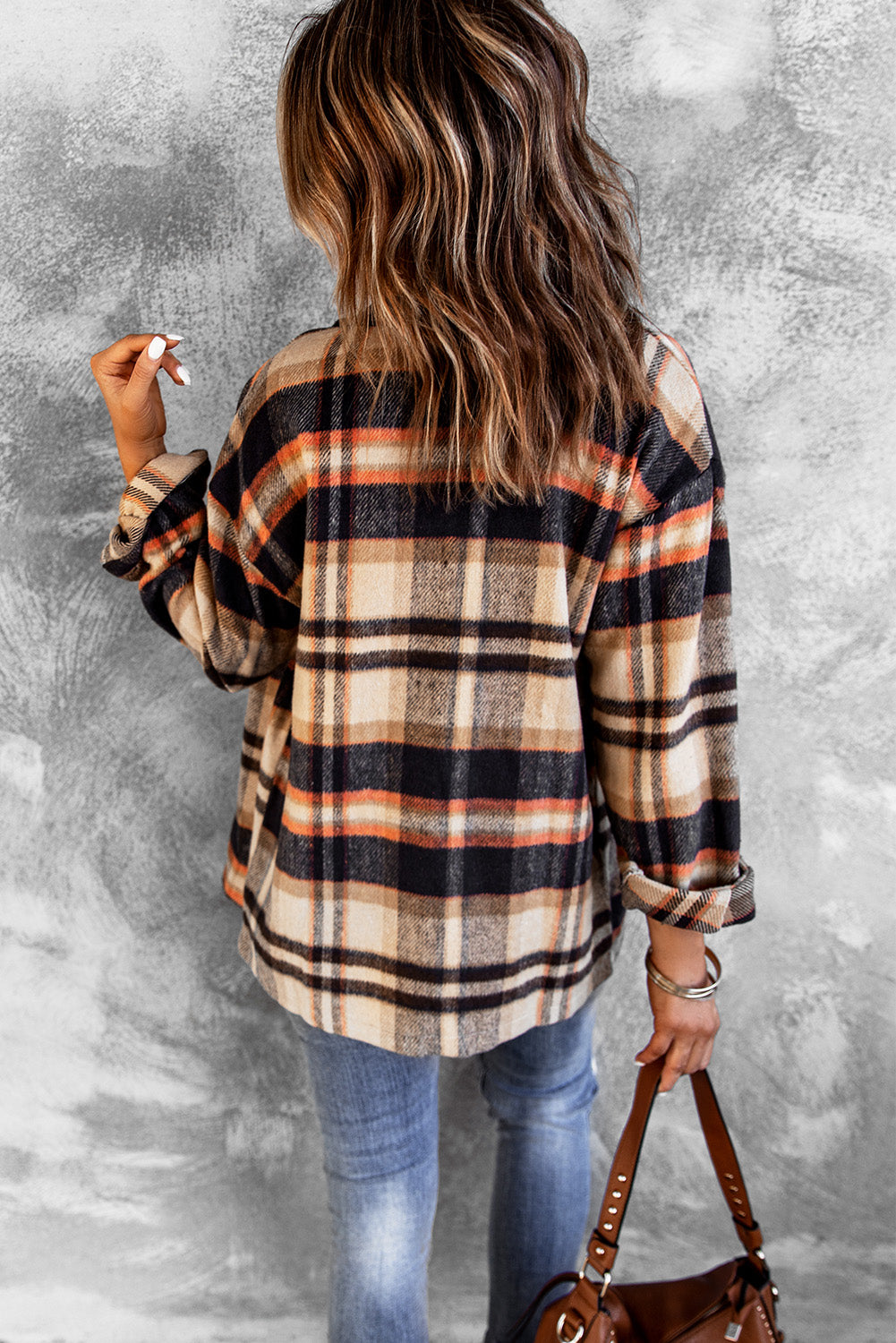 A person wearing a Brown Plaid Button Up Long Sleeve Flannel Shacket layered over a white top with ripped jeans and holding a brown handbag stands against a textured gray wall.