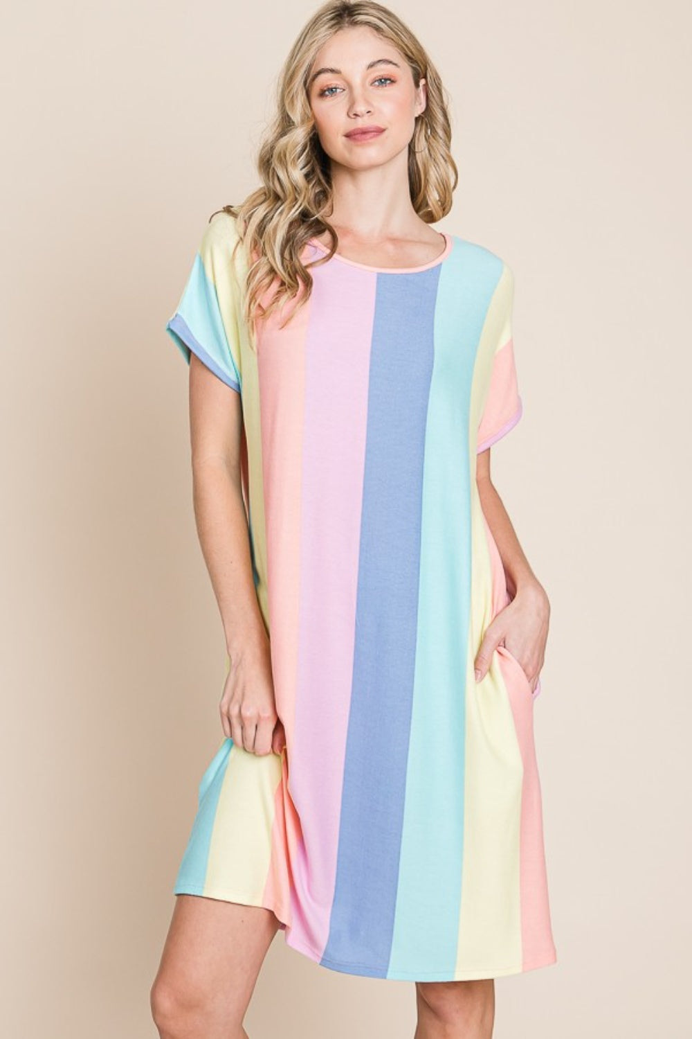 A person stands against a plain background, wearing the BOMBOM Striped Short Sleeve Dress with Pockets, crafted from comfortable fabric and featuring pastel stripes, with hands casually tucked into the convenient pockets.