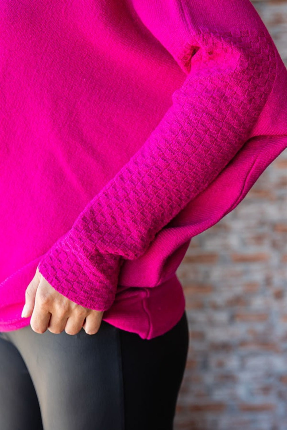 A person with long, blonde hair is standing indoors, smiling, and wearing a bright pink First Love Full Size Asymmetrical Hem Dolman Sleeve Sweater paired with black pants.