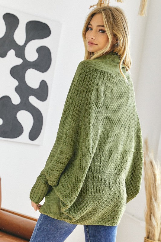 A person stands against a plain background, wearing a Pattern Knit Dolman Sleeve Solid Slouch Cardigan in textured blue over a white sweater with distressed white shorts, hands in cardigan pockets.