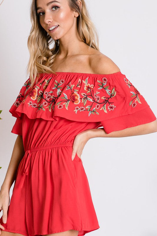 Wearing the stylish Floral Embroidered Off Shoulder Romper, a woman poses gracefully, showcasing its ruffle detail and making it an ideal addition to any summer wardrobe.