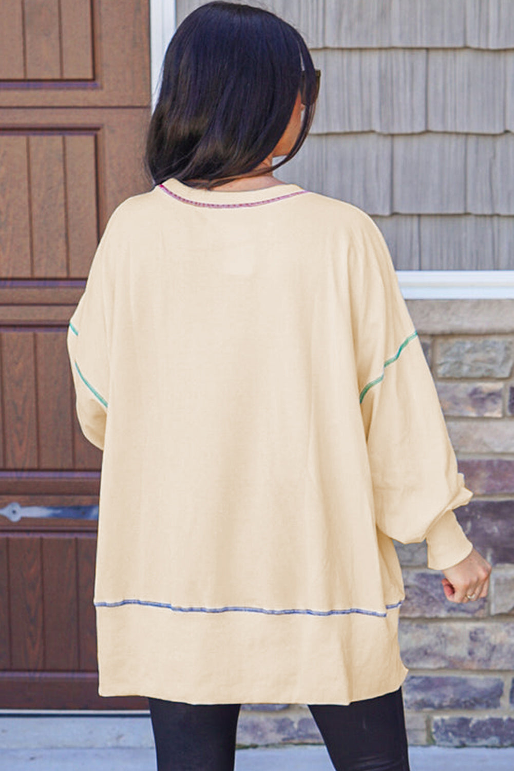 A woman with blonde hair is facing away, wearing a Contrast Stitching Split Baggy Sweatshirt. She stands in a room adorned with plants and wooden furniture in the background.