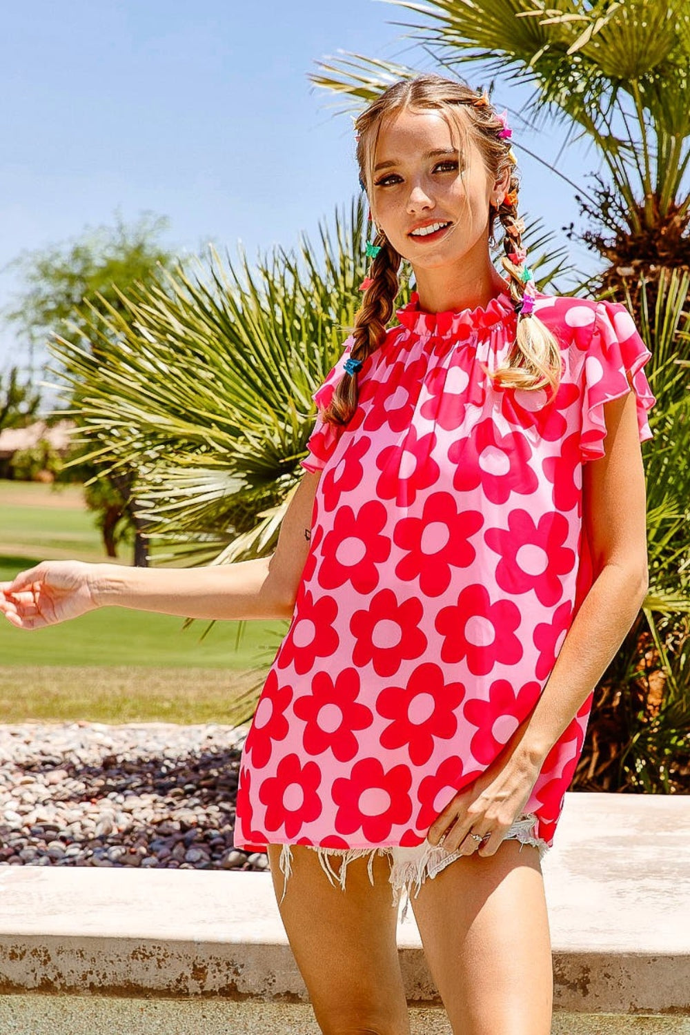 A woman with braided hair wearing a BiBi Floral Contrast Ruffled Top and light denim shorts stands outdoors in front of palm trees on a sunny day.