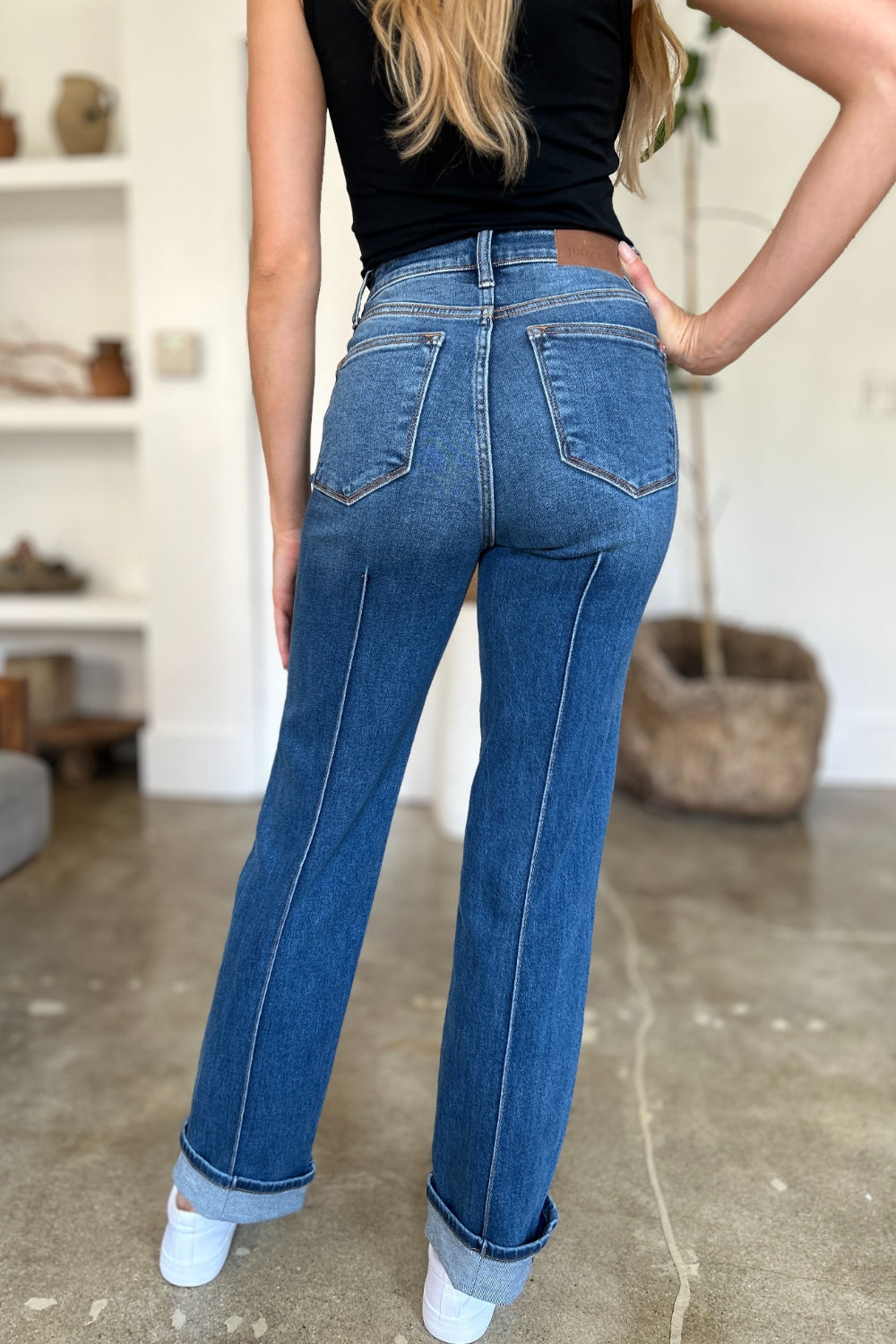 Two women stand side by side indoors, both wearing black tank tops, Judy Blue Full Size High Waist Front Seam Detail Straight Jeans, and white shoes. They are smiling and posing for the camera. A plant and shelves with decorations are in the background.