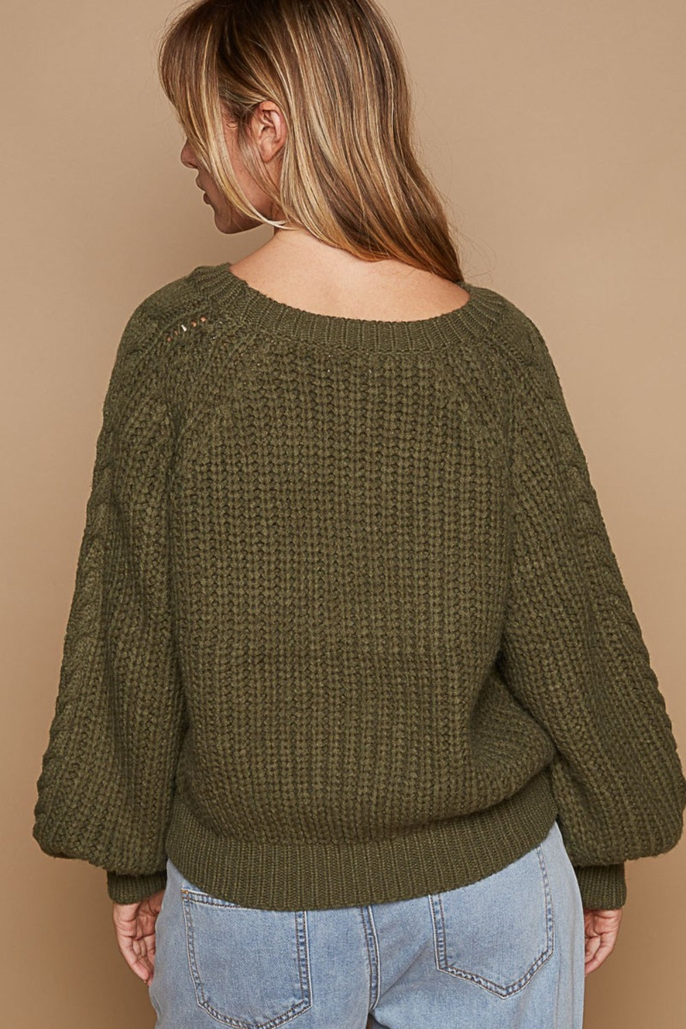 A woman wears the POL Cable Knit Cutout Long Sleeve Sweater in olive-green, showcasing a subtle shoulder keyhole, beautifully matched with light blue jeans adorned with floral embroidery, all set against a beige background.