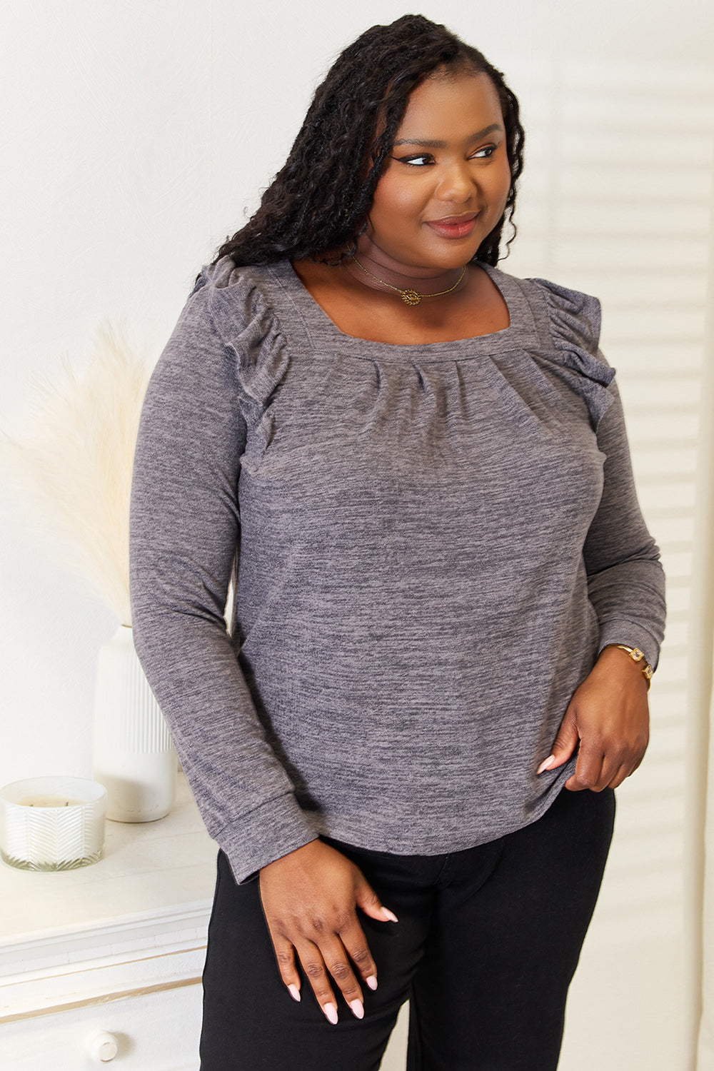 A woman wearing a Double Take Square Neck Ruffle Shoulder Long Sleeve T-Shirt in pink and blue jeans stands with one hand in her pocket, smiling. She has long curly hair and wears a necklace and bracelet. She stands in a room with white decor.