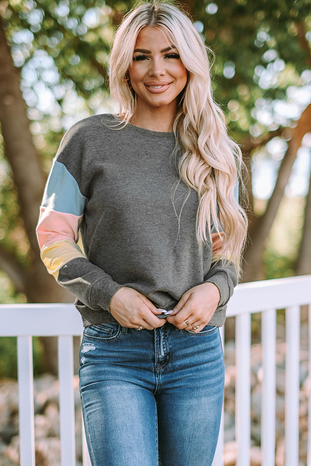 A person with long blonde hair wearing a stylish Gray Colorblock Long Sleeve Pullover Sweatshirt and blue jeans stands outdoors near a white fence.