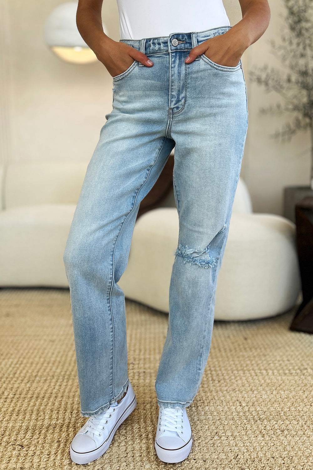 A person standing on a beige carpet, wearing Judy Blue Full Size High Waist Distressed Straight Jeans in light blue, a white top, and white sneakers.