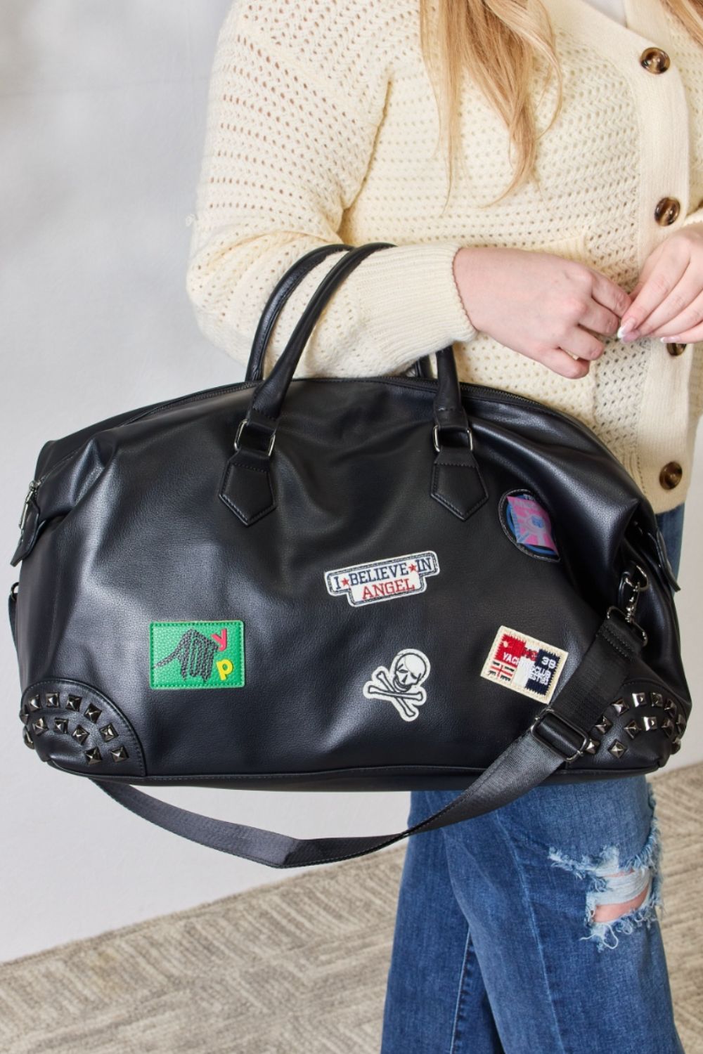 A person holding the SHOMICO Rivet Detail Patch Handbag, a black PU leather duffel bag with a colorful patchwork design, wearing a cream sweater and blue jeans.