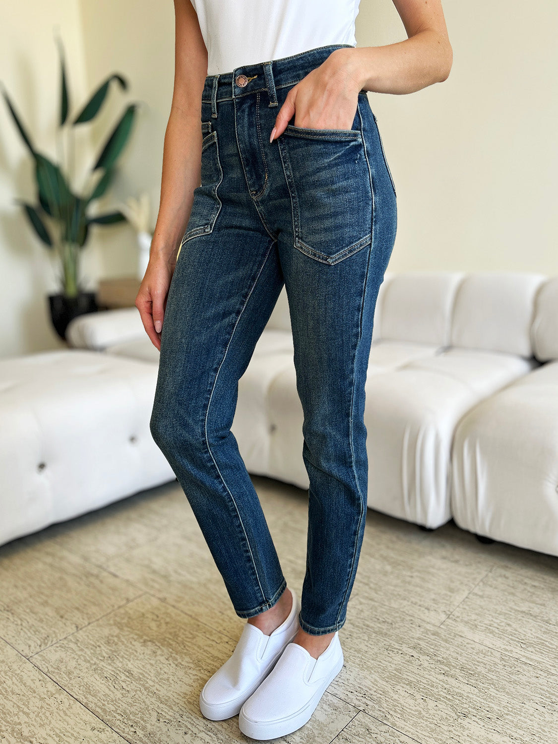 A person wearing Judy Blue Full Size High Waist Skinny Jeans made of stretchy denim fabric stands in a room with light-colored furniture and wooden flooring, paired perfectly with white slip-on shoes.