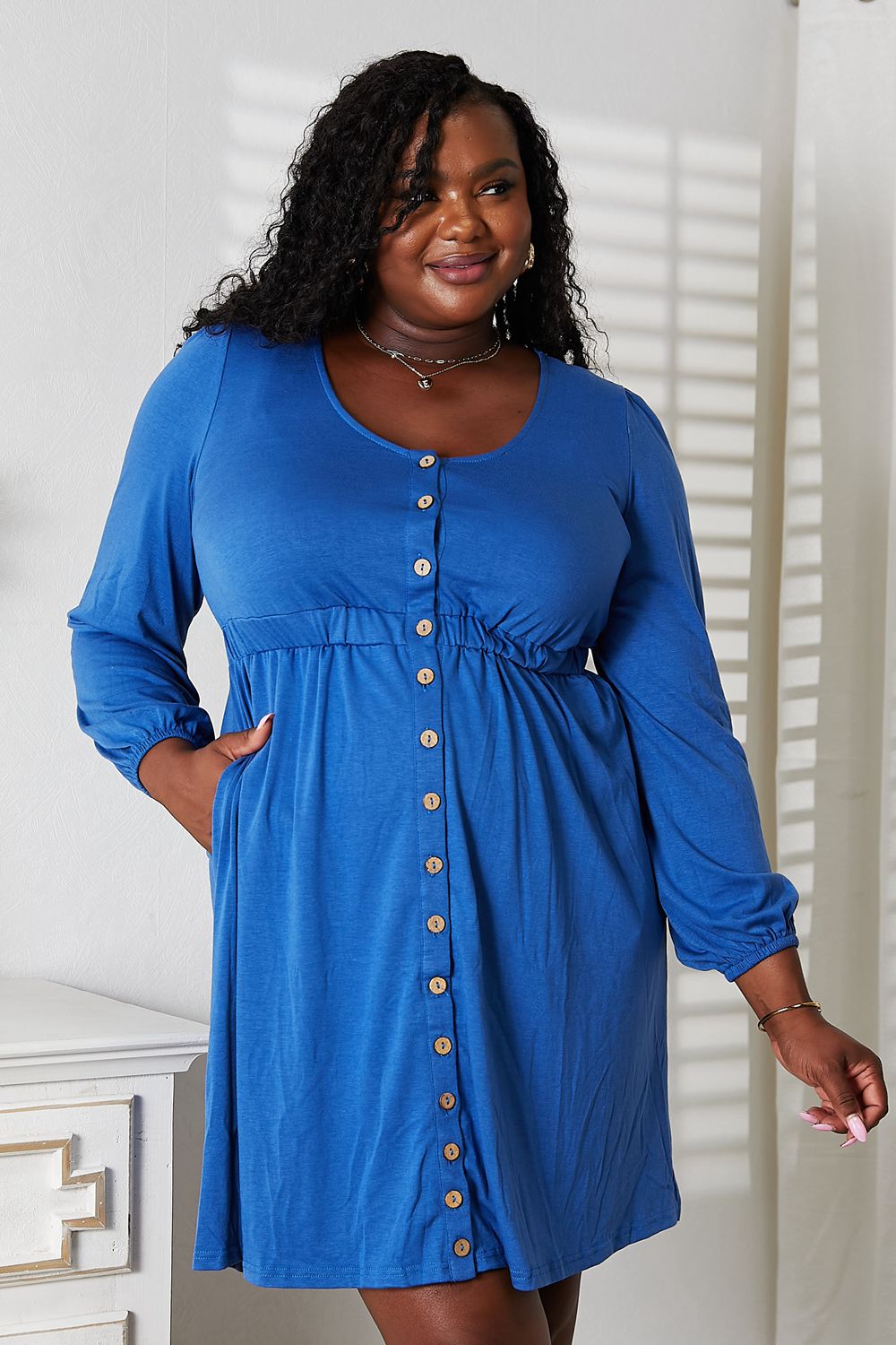 A woman poses indoors, smiling and holding out the skirt of a blue Double Take Scoop Neck Empire Waist Long Sleeve Magic Dress. She has curly hair and stands in front of a white wall with a partially visible piece of furniture.