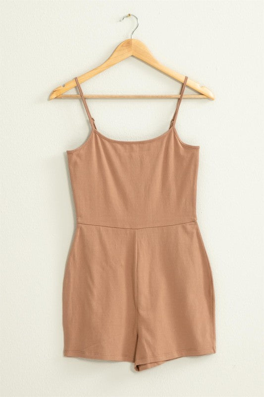 The Loving Me Fitted Sleeveless Romper, featuring a fitted bodice and spaghetti straps in black, hangs gracefully on a wooden hanger against a white wall.