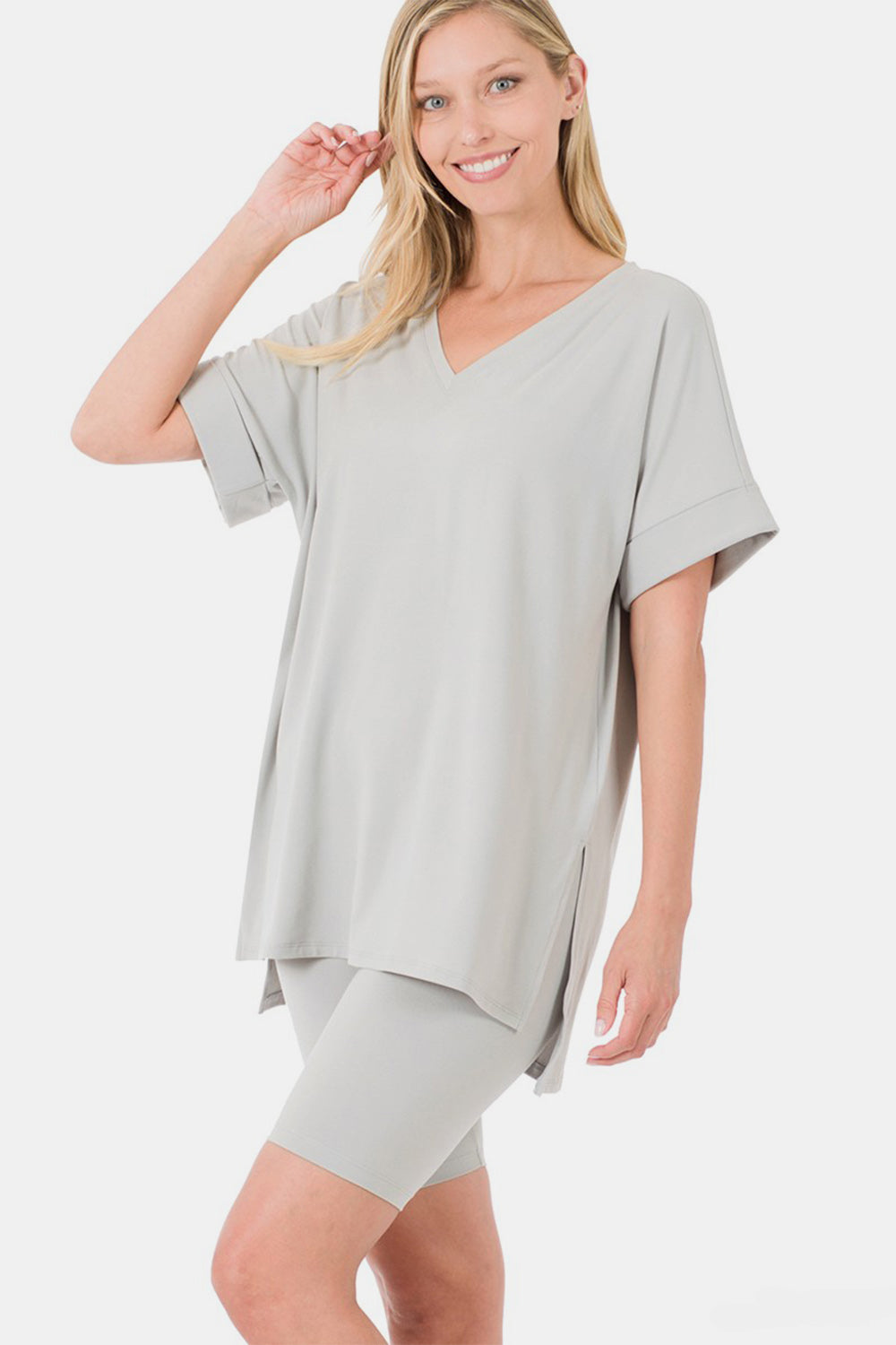 A woman is wearing the Zenana Full Size V-Neck Short Sleeve Slit T-Shirt and Shorts Set in a light gray color. The loose-fitting, highly stretchy outfit features a V-neck t-shirt with short sleeves and matching knee-length shorts. She is smiling with her hand raised near her head.