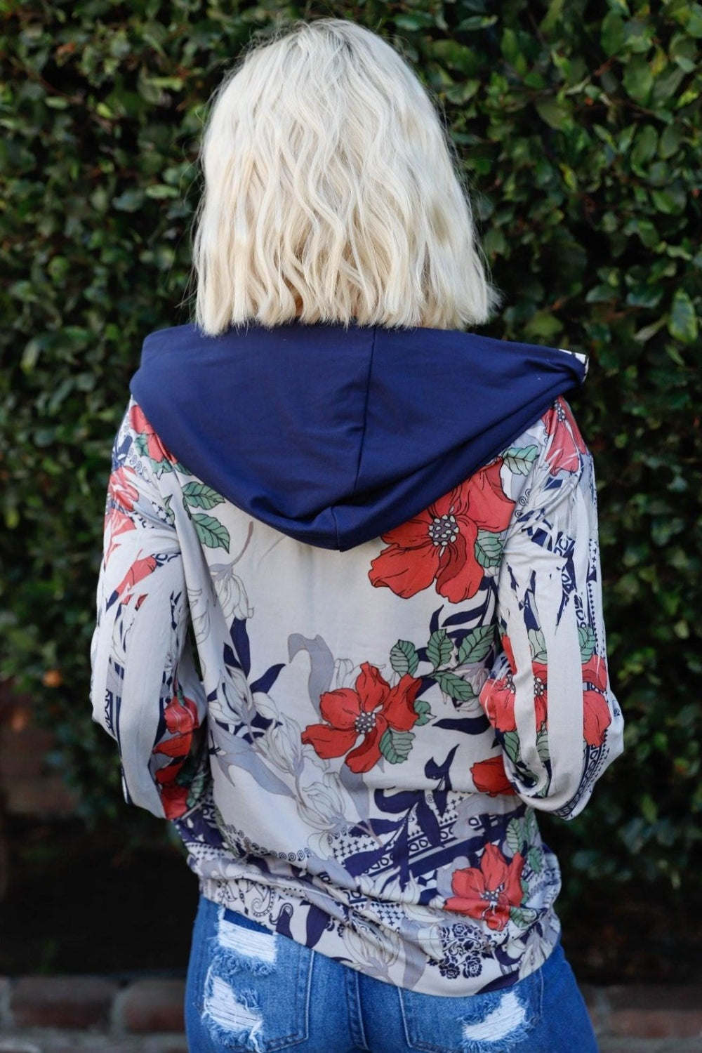 A woman stands outdoors wearing the Blumin Apparel Full Size Floral Zip-Up Hoodie with her hands in the pockets. A leafy green hedge is in the background, adding to the casual chic vibe.