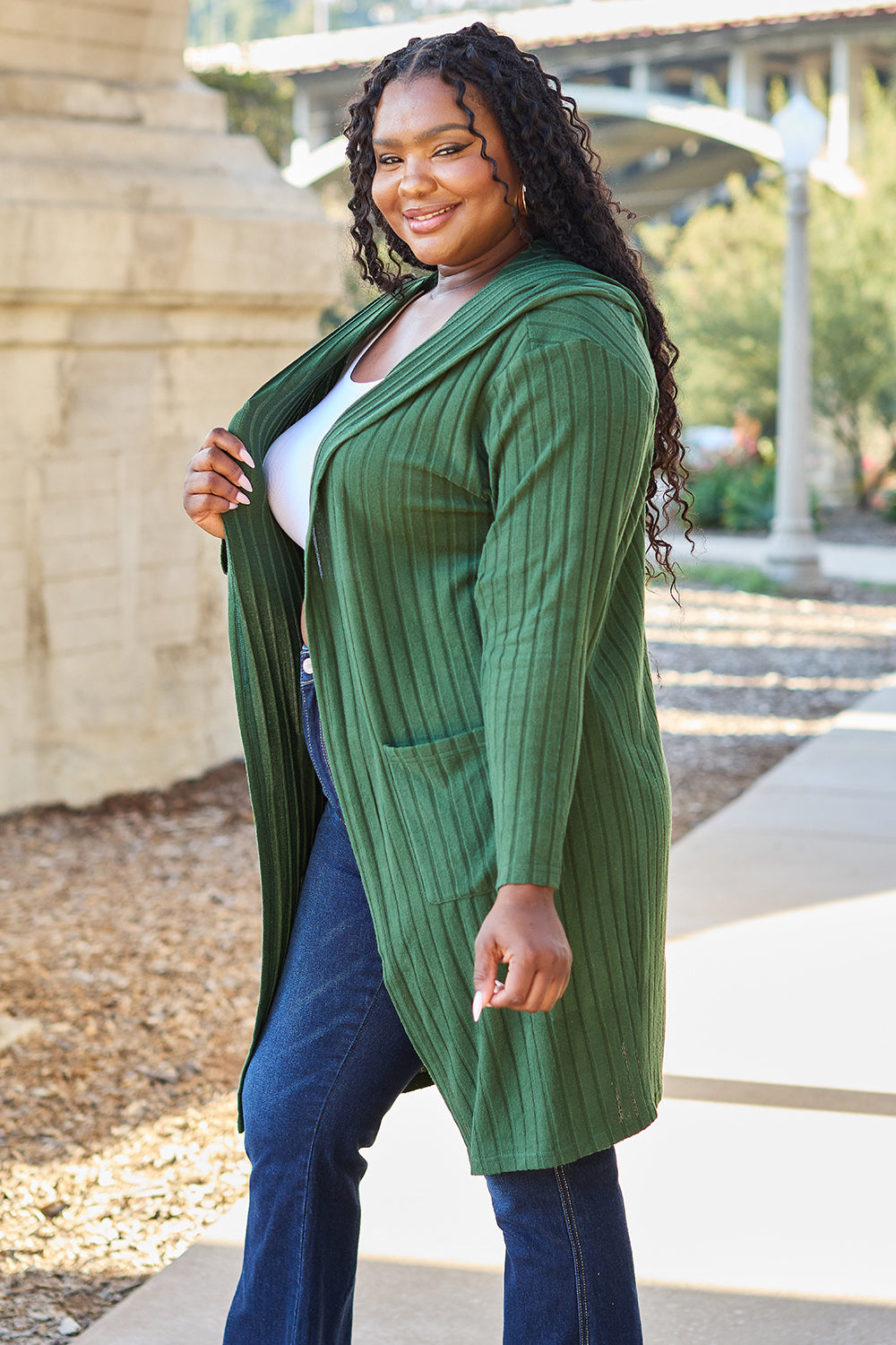 A woman wearing the Basic Bae Full Size Hooded Sweater Cardigan in black, paired with a white top and khaki pants, stands in front of a light background accented by a leafy plant on her left. With one hand tucked into her pocket, she gazes to her right.