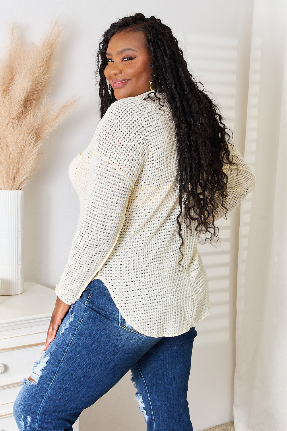 A woman with long, curly hair wearing the Culture Code Full Size Scoop Neck Patch Pocket Top and ripped jeans stands indoors near a white dresser and pampas grass decor.