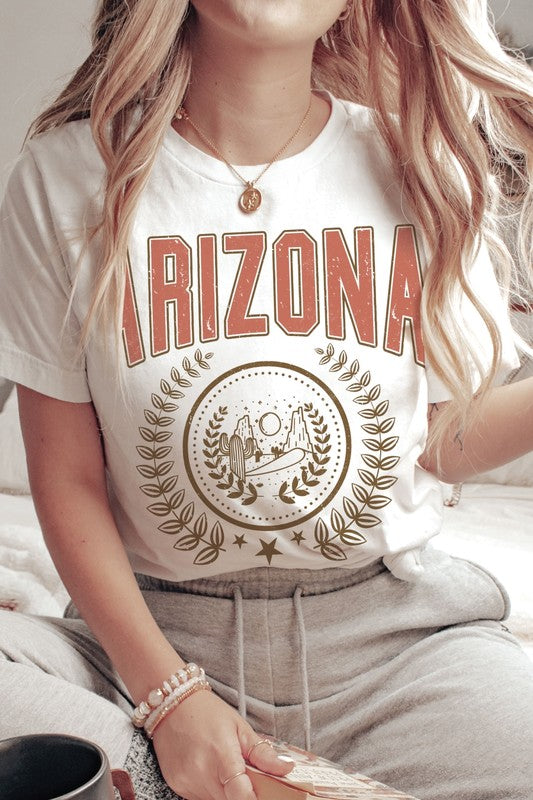 A person wearing a light gray ARIZONA Graphic Tee that showcases a desert scene with cacti and a sun, encircled by a laurel wreath design. This tee is made from 100% cotton and is available in unisex sizing.