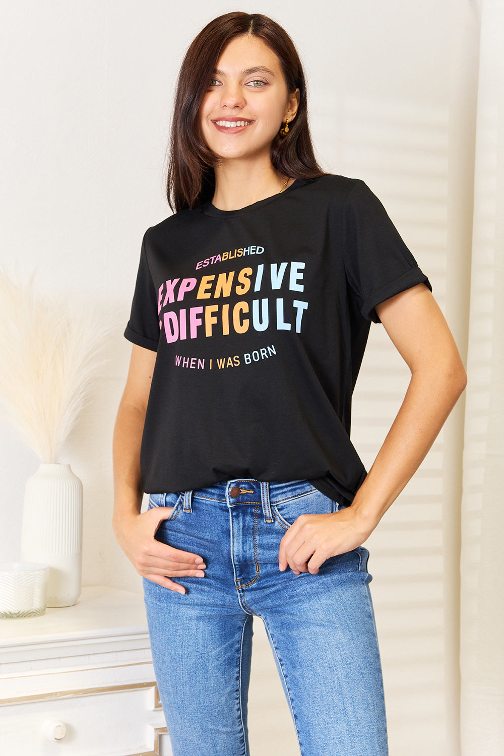 A woman with long braided hair, wearing the Simply Love Slogan Graphic Cuffed Sleeve T-Shirt that says "Established Expensive & Difficult When I Was Born," stands with one hand in her pocket. The trendy cuffed sleeves of her shirt add an extra touch of style.