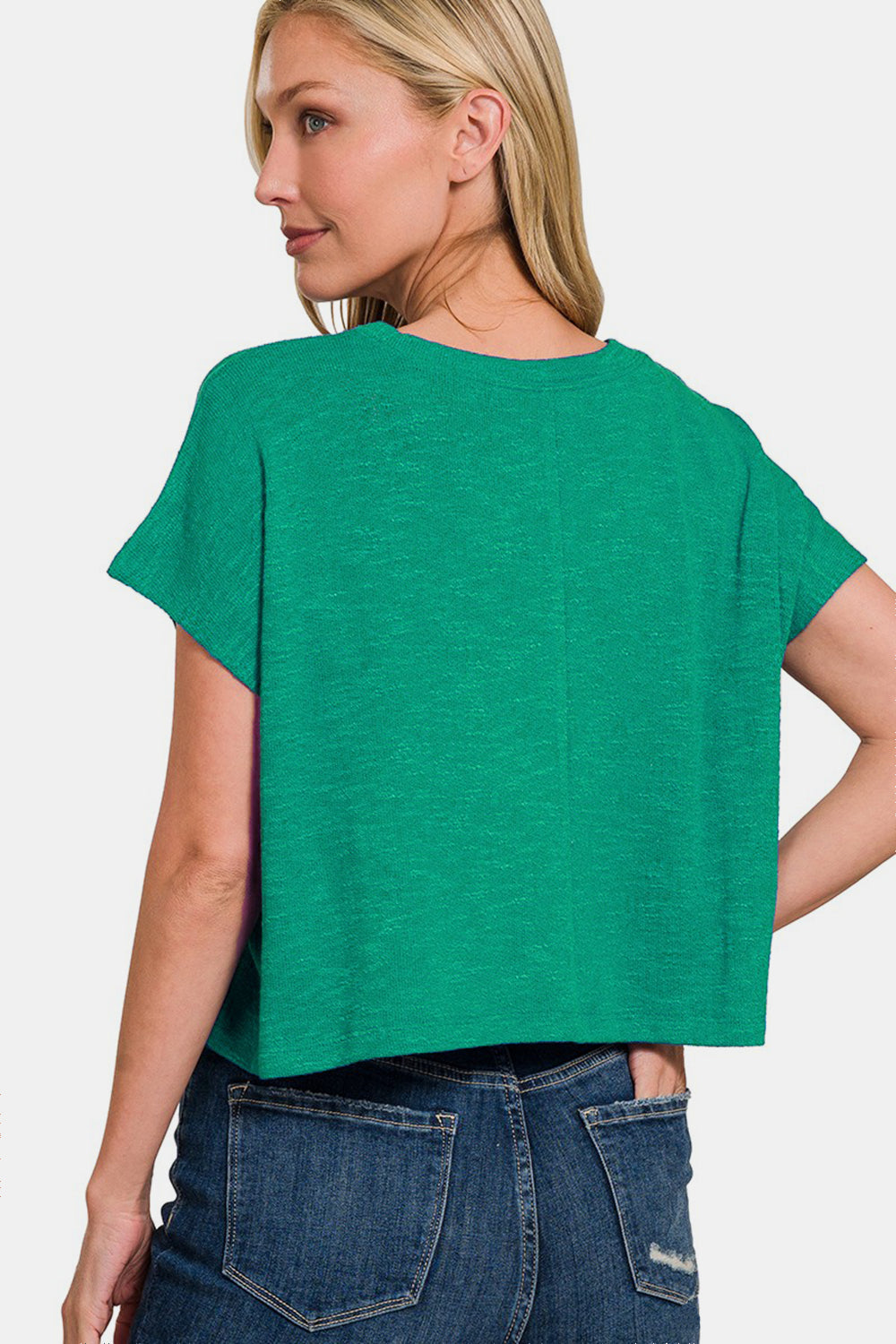 A person with long, blond hair is wearing a green Zenana Round Neck Short Sleeve T-Shirt and blue jeans, with their left hand in a pocket, smiling slightly and looking to the side.